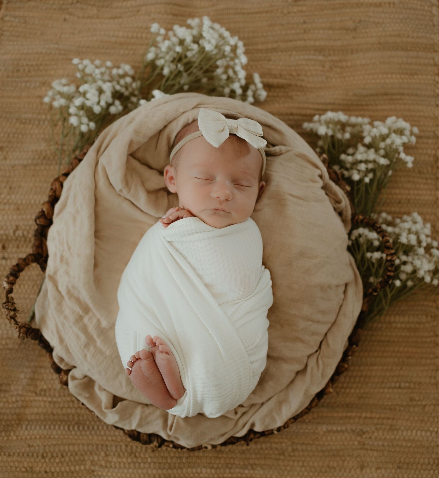 Newborn baby wrapped in white swaddle sleeping on beige blanket with baby's breath flowers.