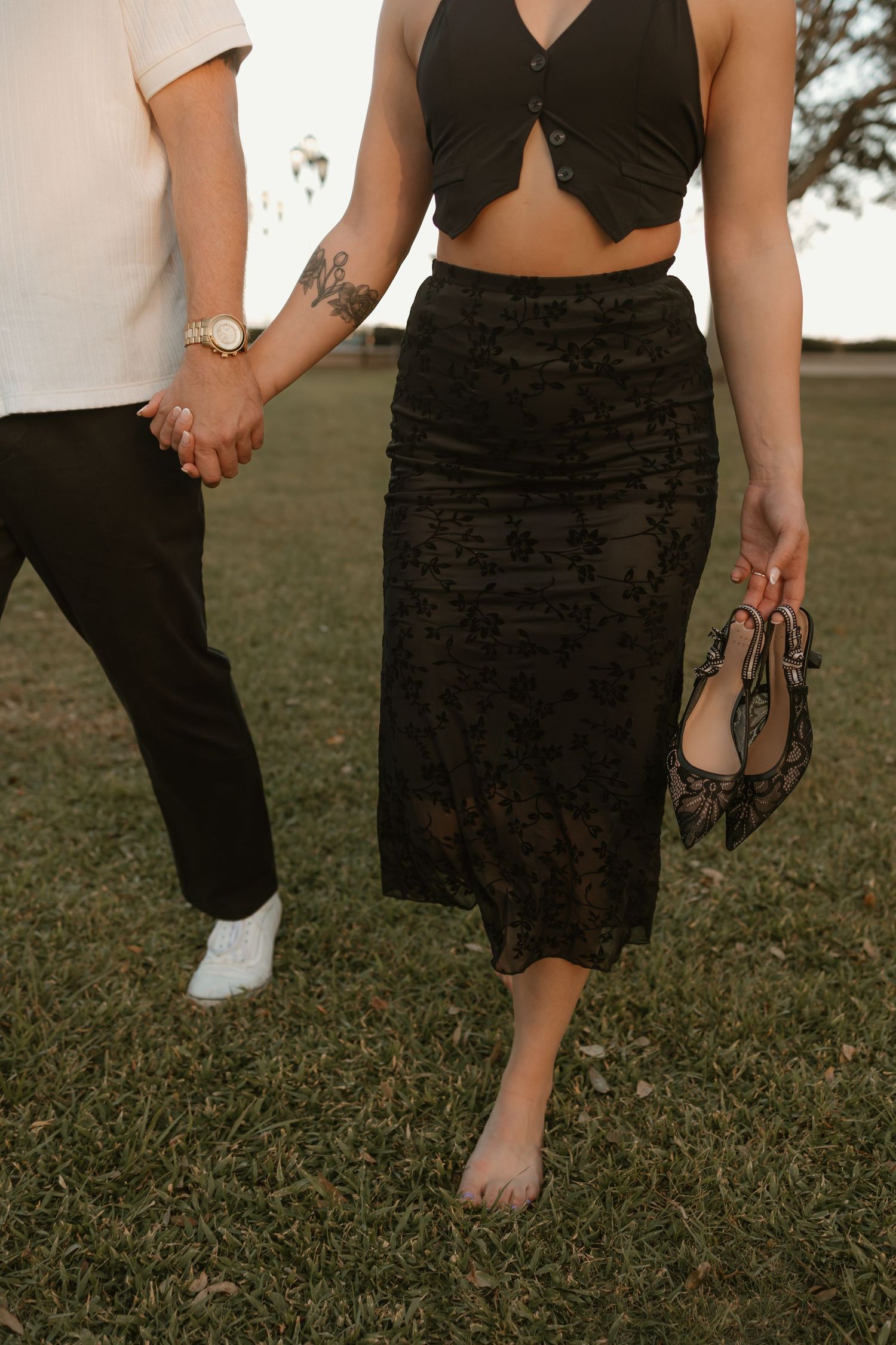 Lower body shot of couple walking barefoot on grass holding hands.