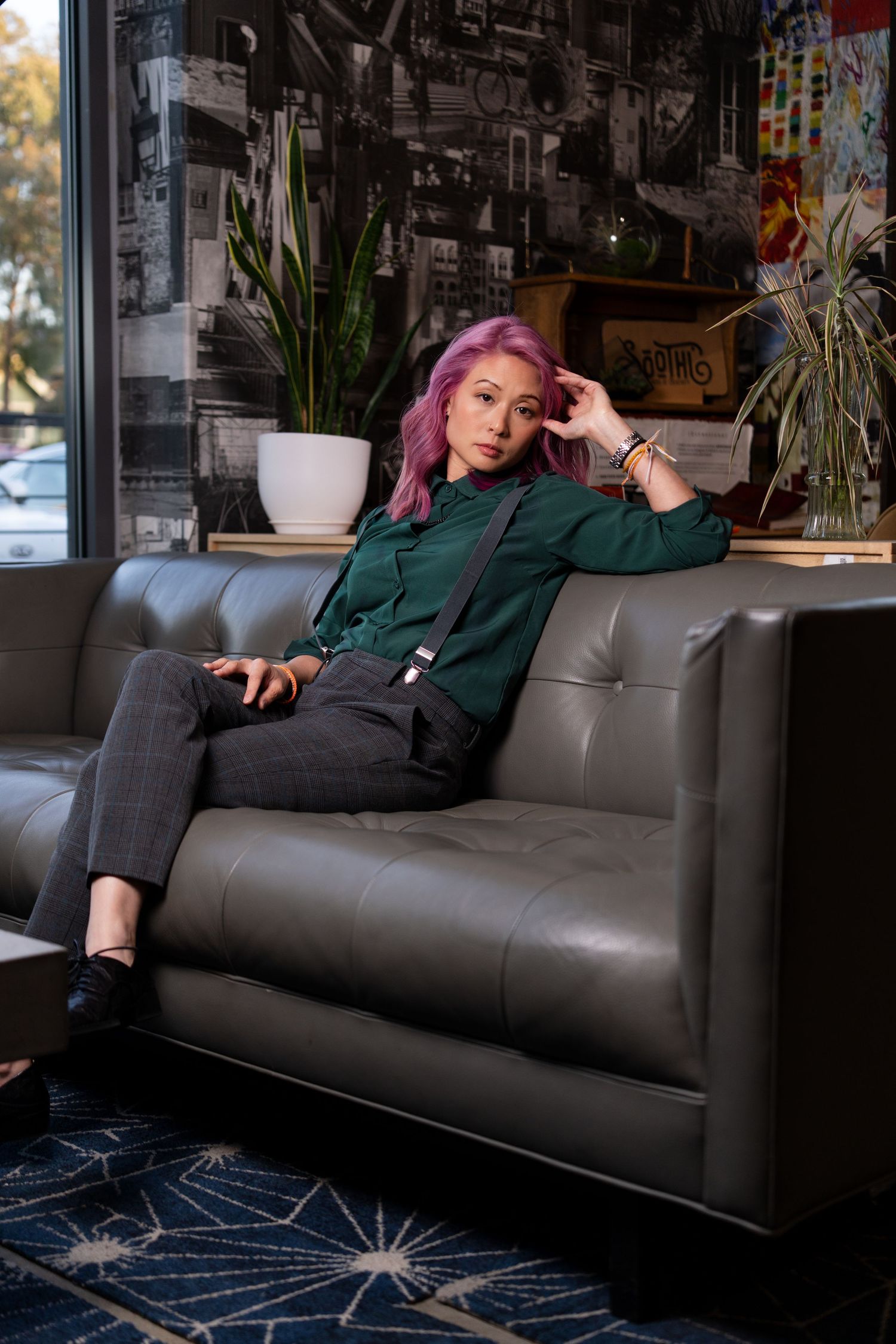 A person with purple hair relaxes on a dark leather couch in a dimly lit room with indoor plants in the background.
