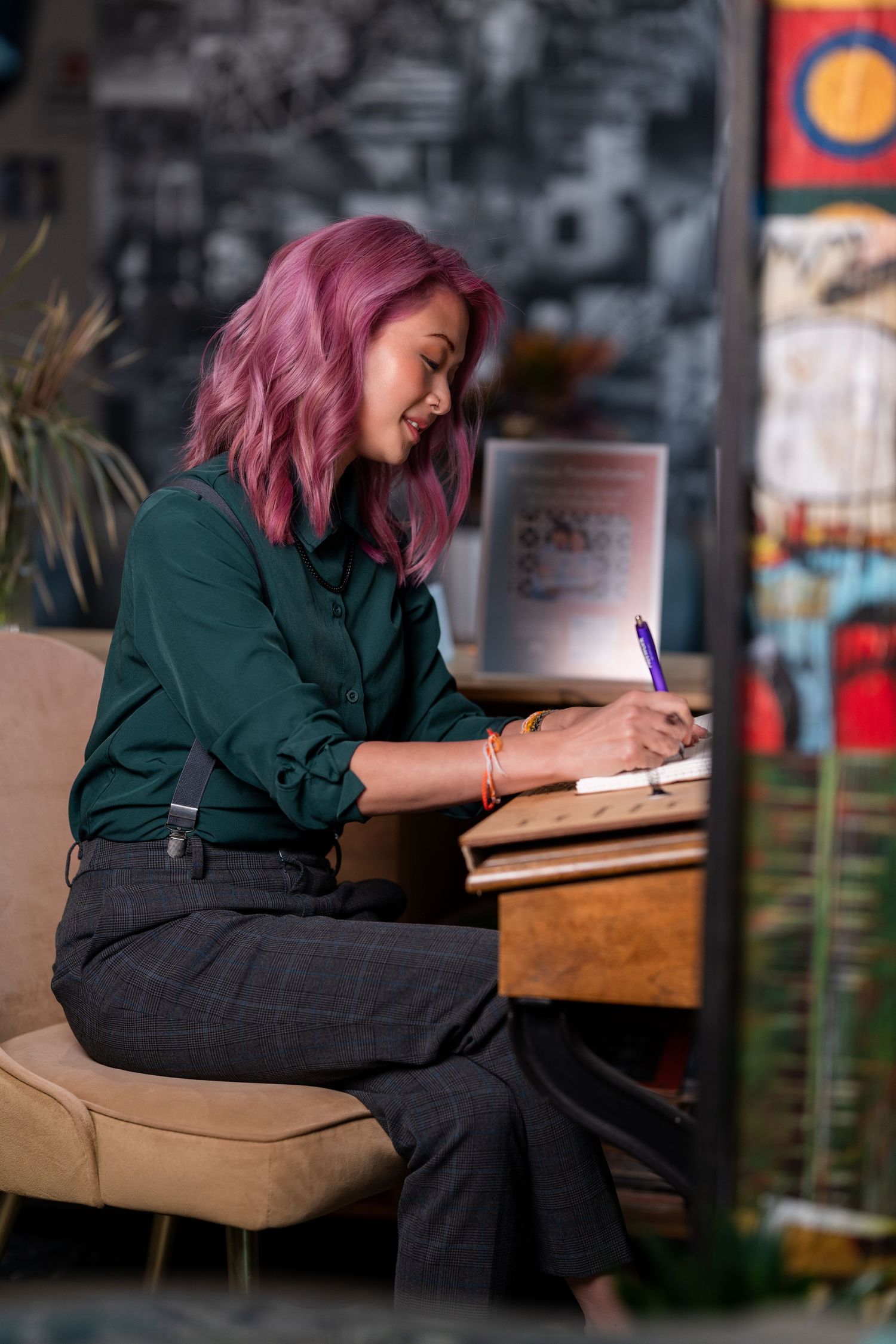 Person with pink hair wearing dark clothing writes in notebook while sitting at table in dimly lit cafe setting.