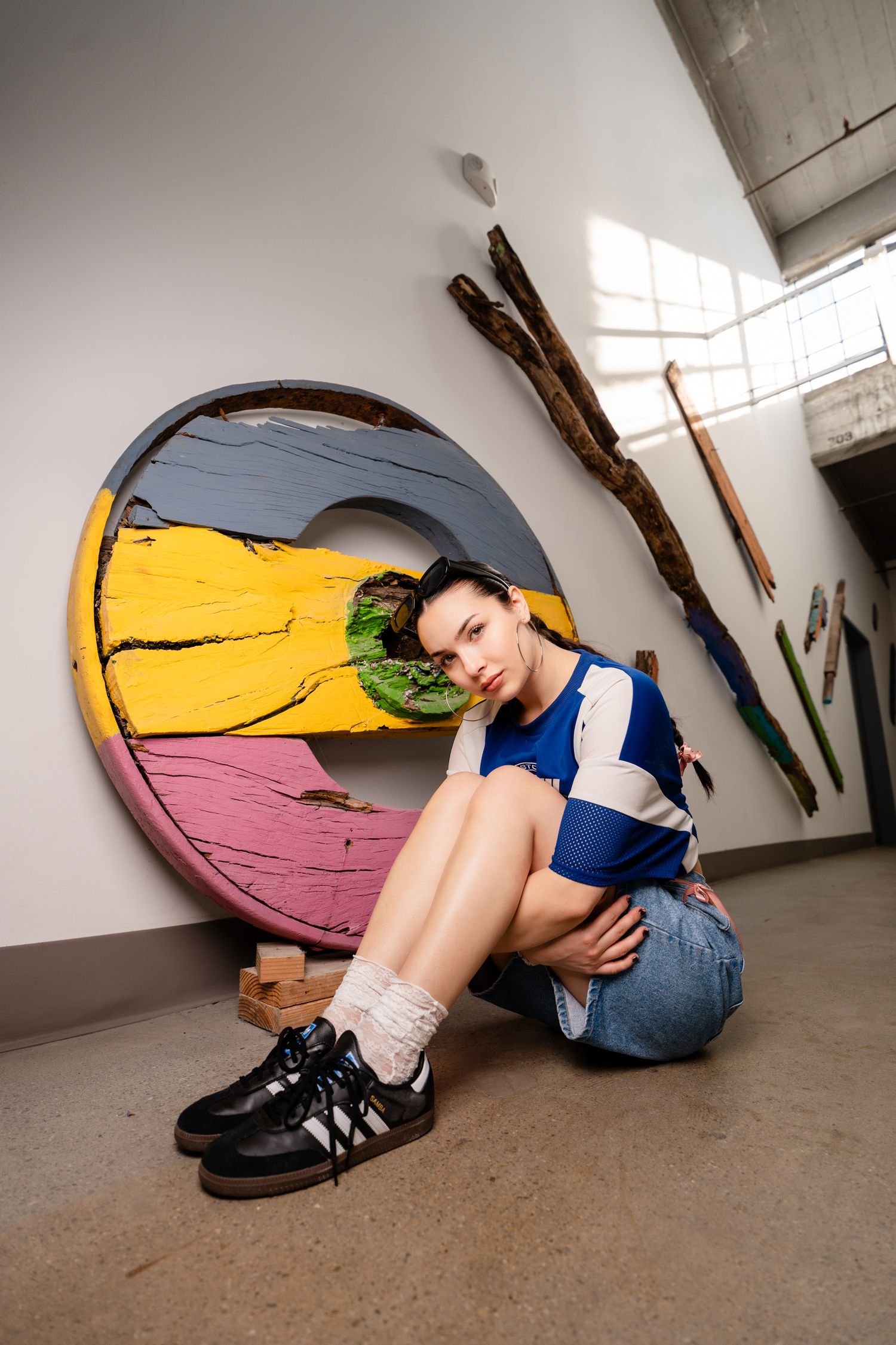 Person in blue shirt and jeans curled up against wall with colorful circular artwork in industrial space.