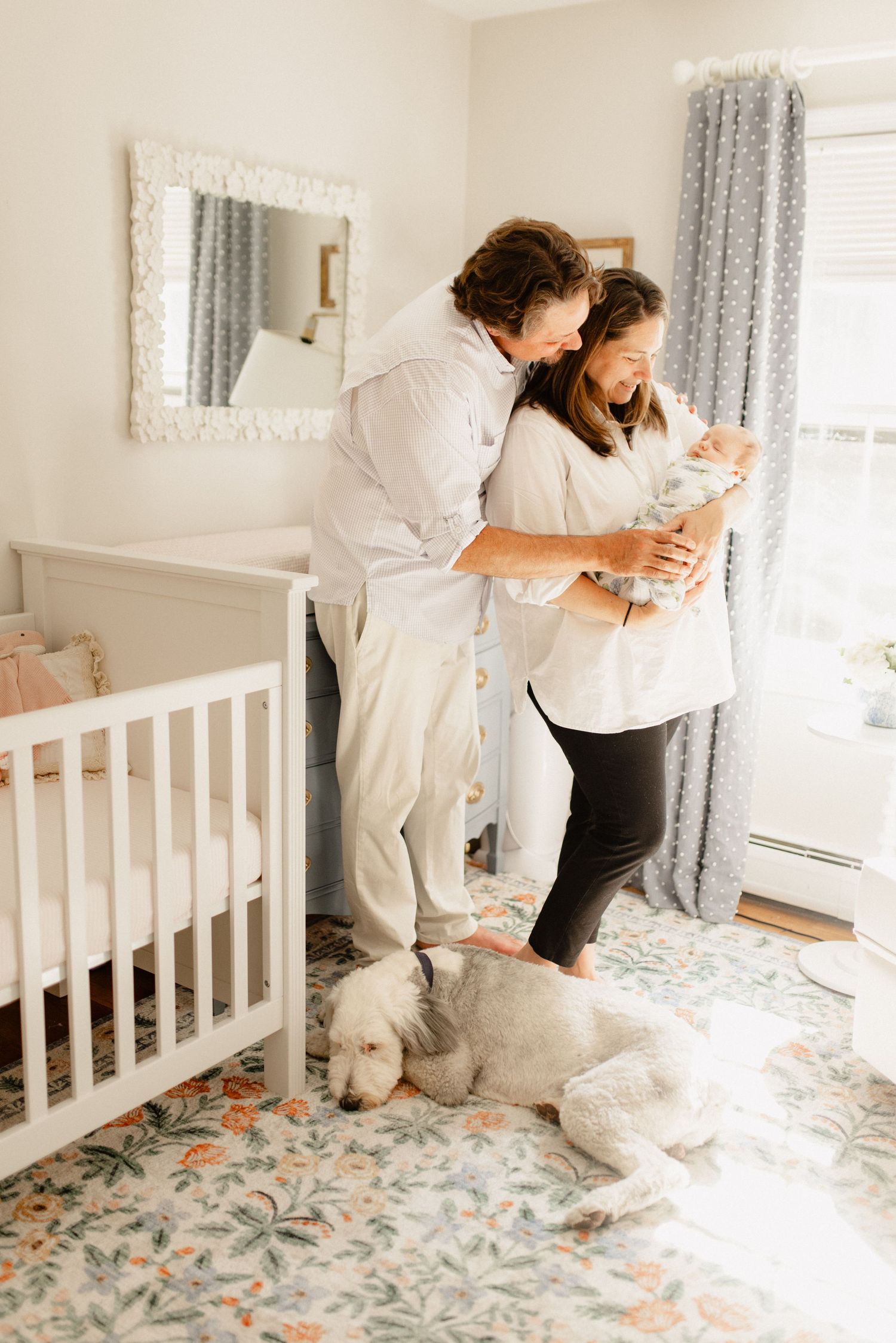 A family cuddles with their newborn in a nursery with a white crib and floral rug.