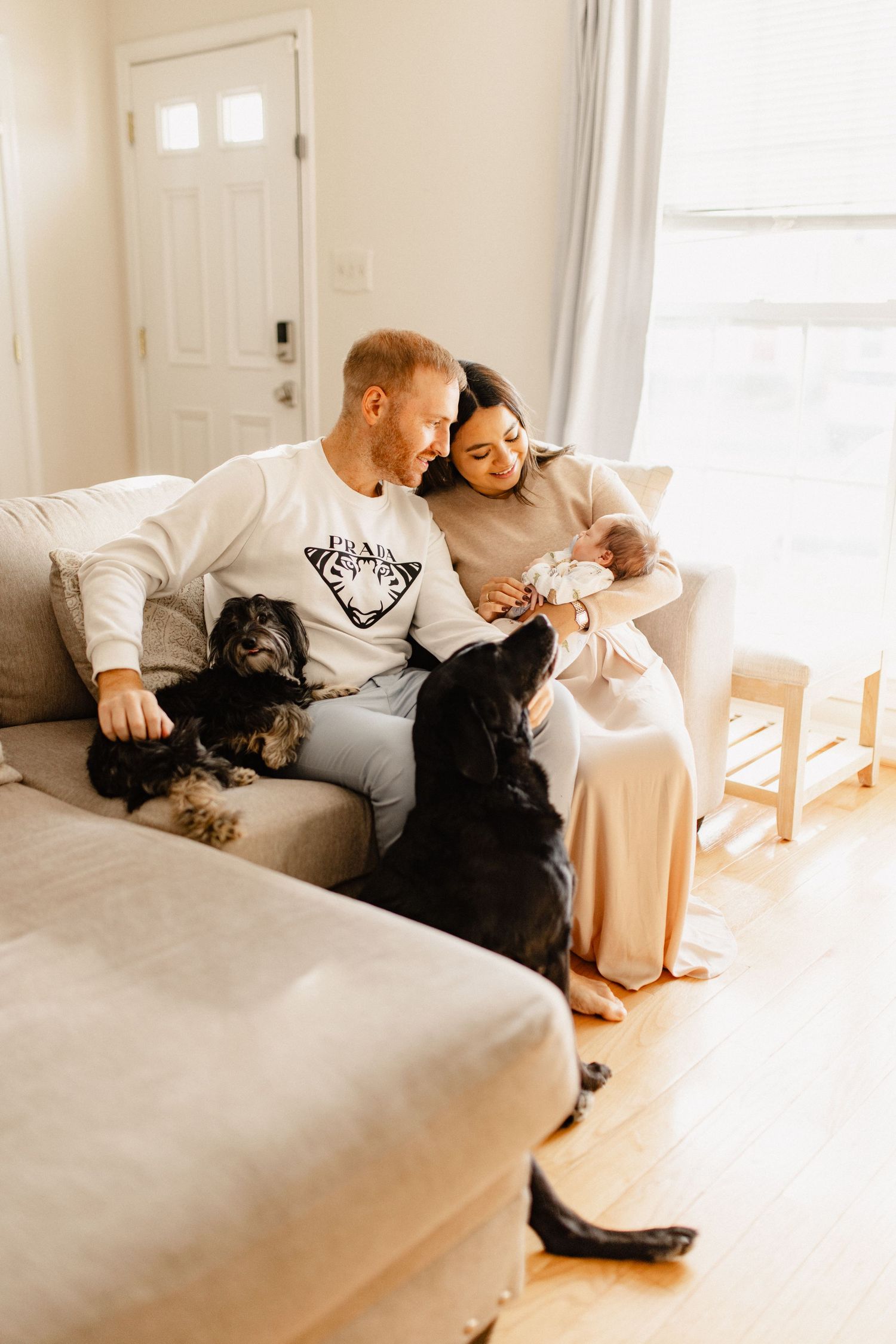 A family relaxes together on a gray couch with two black dogs while holding a baby in a bright living room.