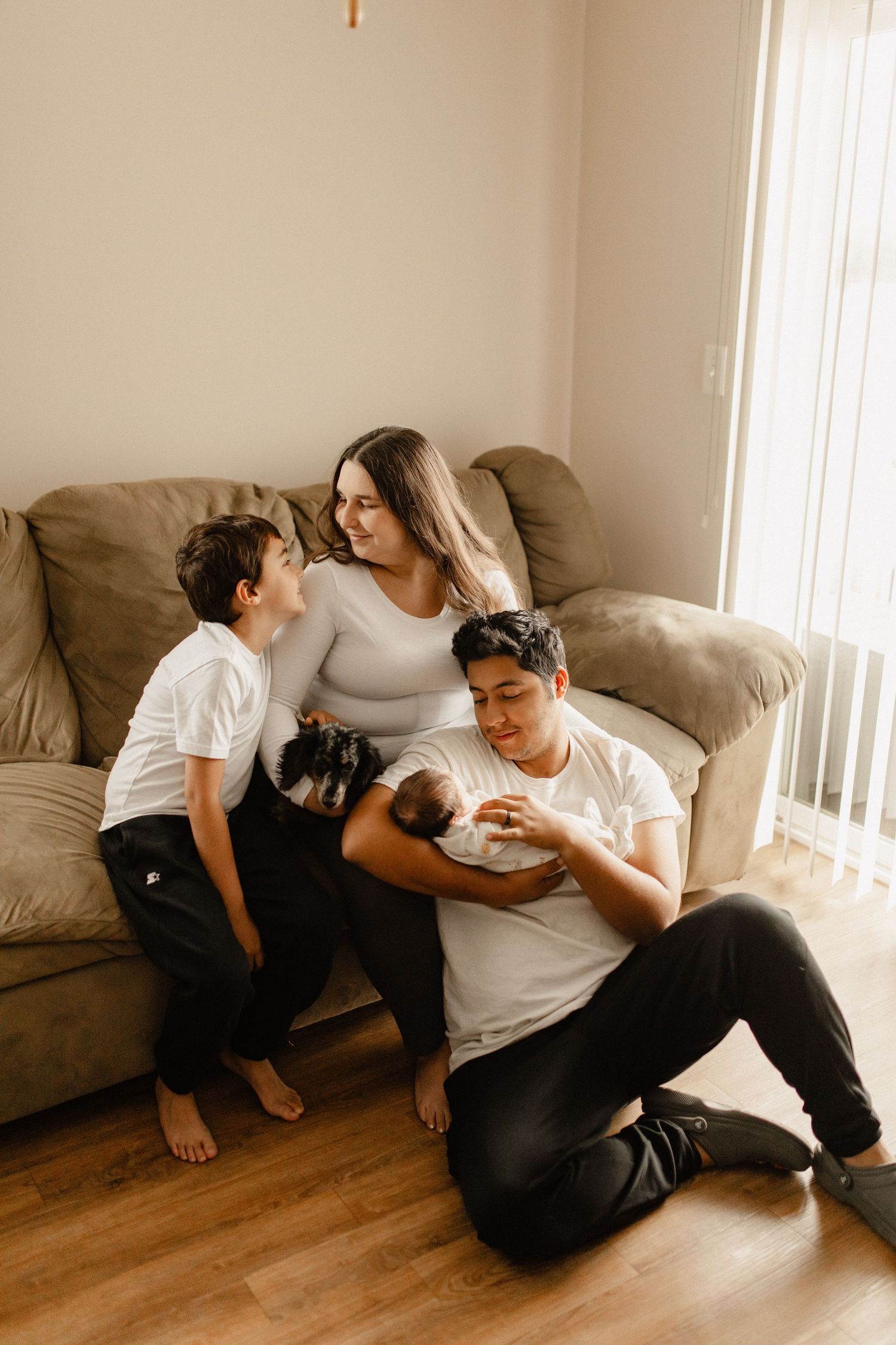 A family lounges together on their brown couch with their cat in a cozy living room.