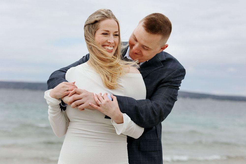 Couple embracing on beach in wedding attire, bride in white gown laughing as groom hugs her from behind near ocean.