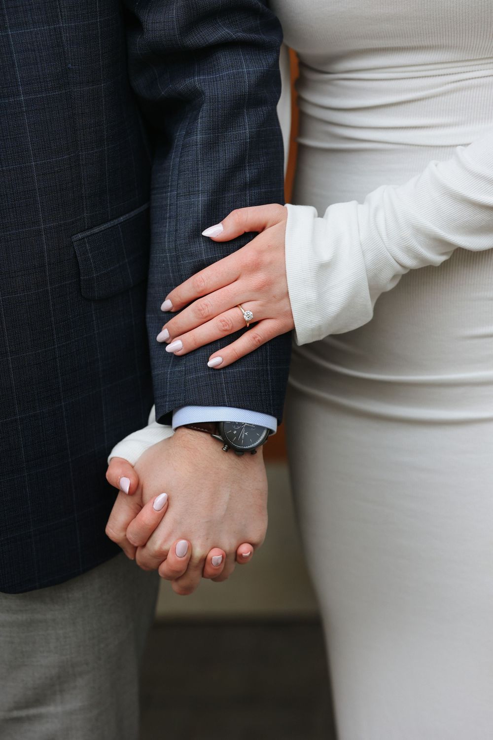 Couple holding hands showing engagement ring and watch, woman in white dress and man in navy suit.