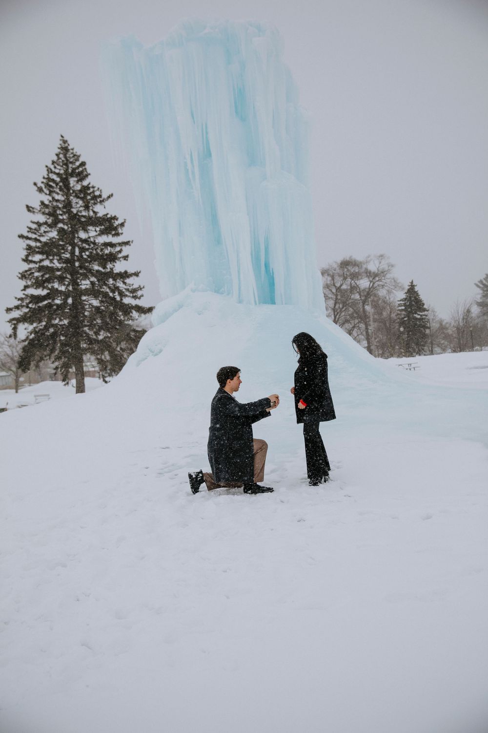 A snowy proposal scene near an ice formation with evergreen trees in the background.