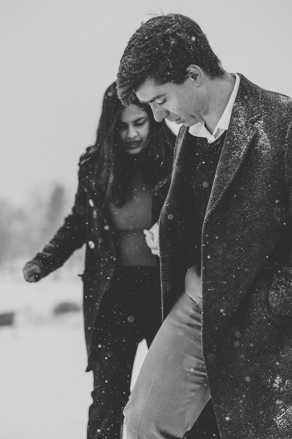 A couple in winter coats laughs together as snow falls around them.