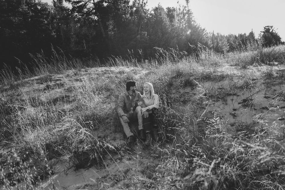 A black and white photograph shows two figures sitting together in a grassy hillside field on a sunny day.