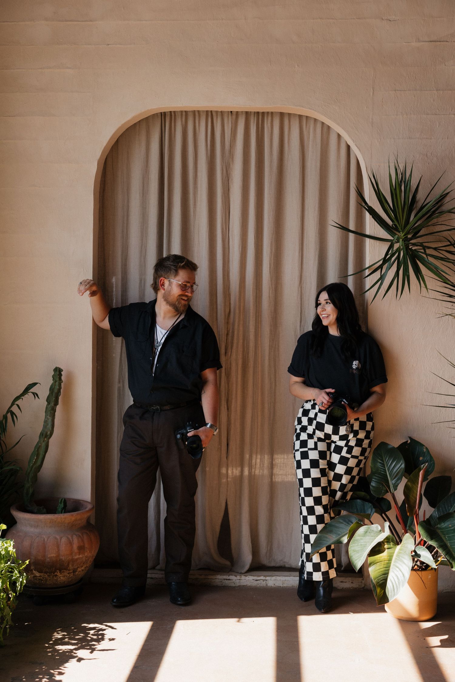 Two photographers in black outfits stand against a beige wall with an arched doorway and potted plants.