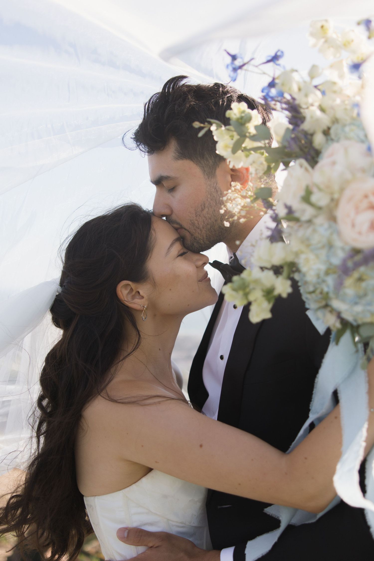 Romantic veil photo of groom kissing bride’s head at Padre Serra Cross in Ventura on a windy day