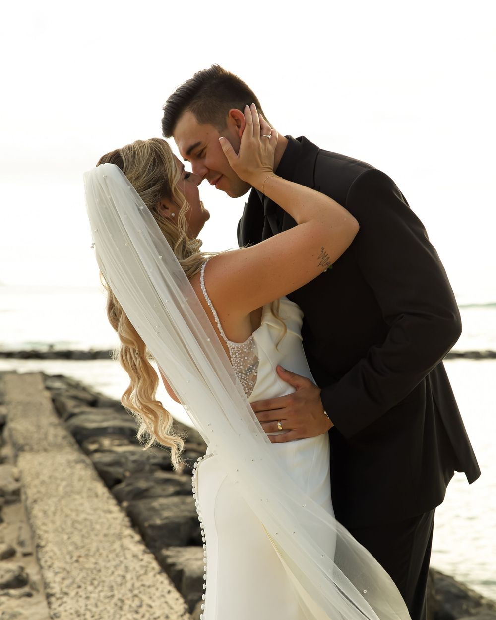 A romantic dip style kiss between newlyweds on a scenic beach with rock jetty in the background.