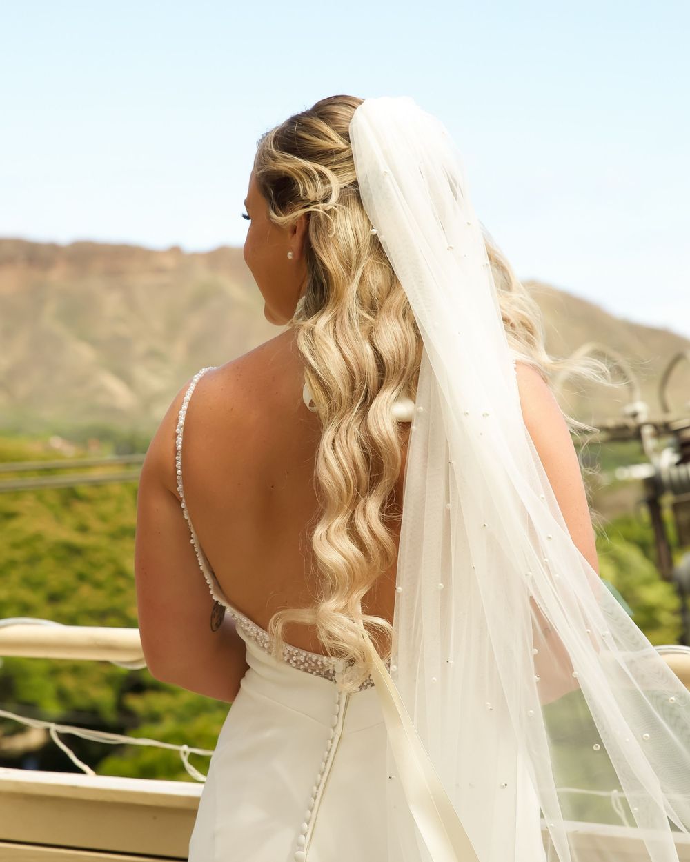 A blonde bride in an elegant white dress and flowing veil stands on a balcony overlooking scenic mountains.