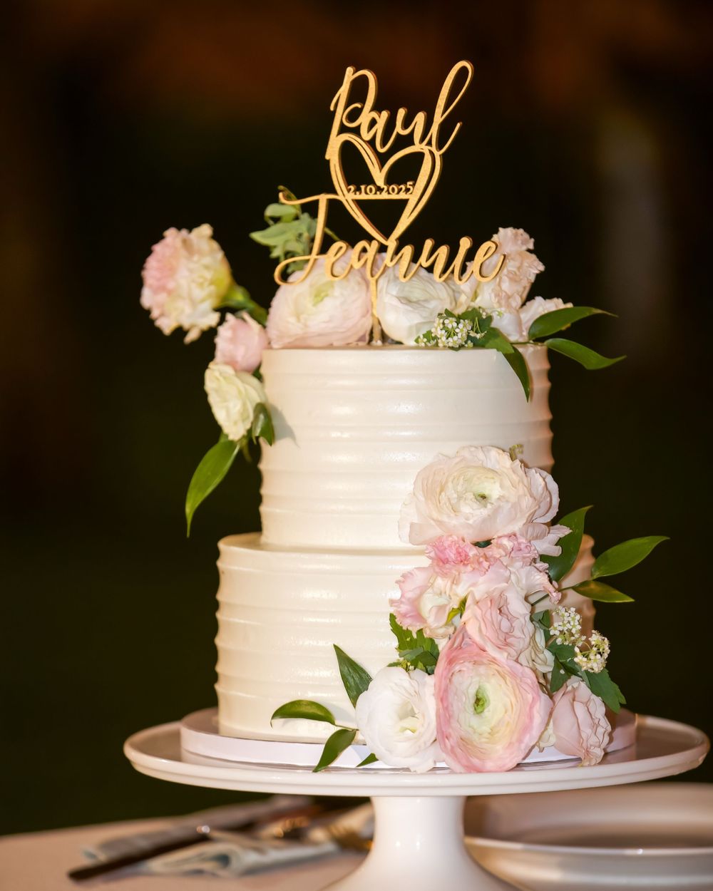 Elegant three-tiered white wedding cake decorated with pink ranunculus flowers and greenery on a white cake stand.