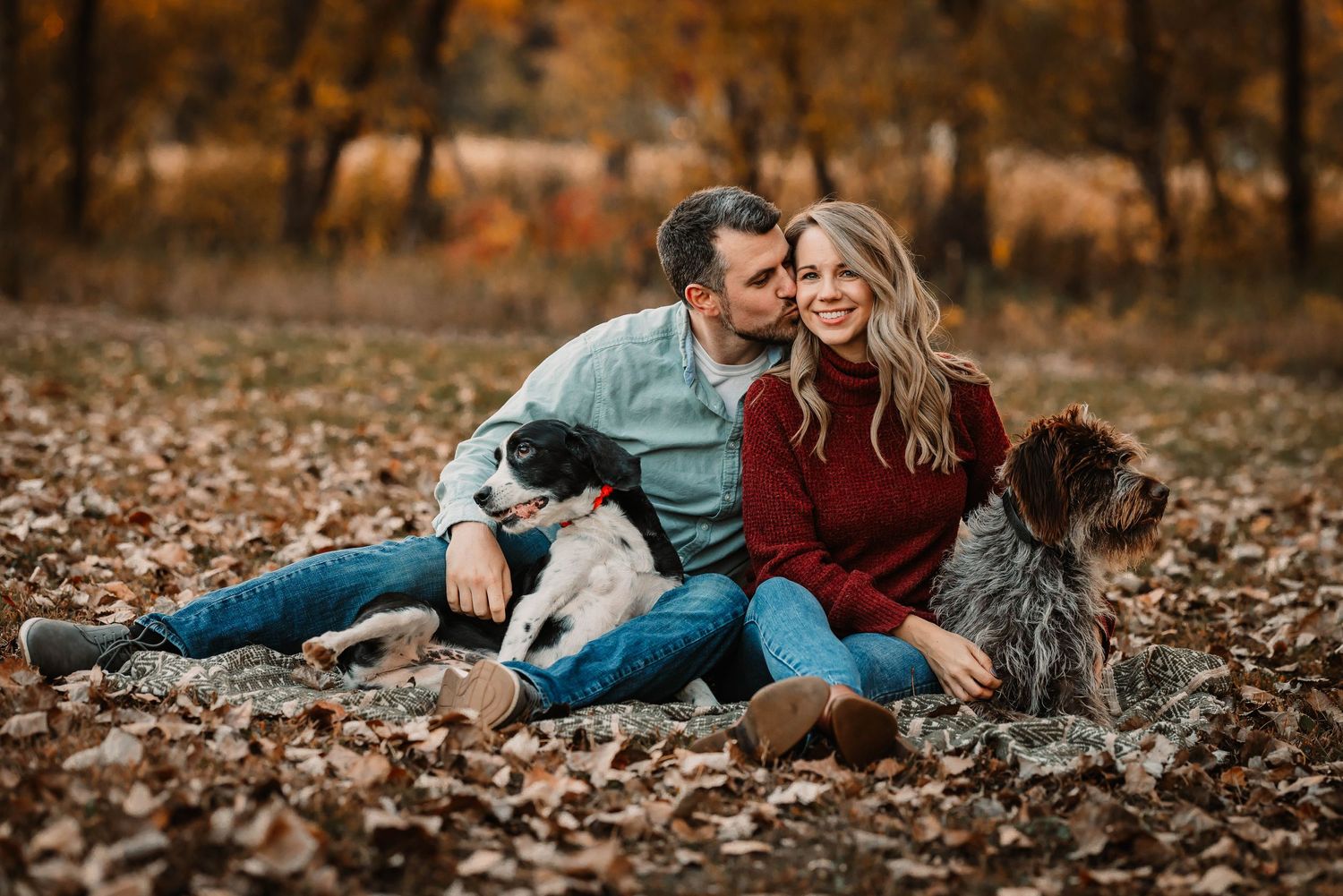 A couple sits with their dog in fallen autumn leaves wearing casual clothing.