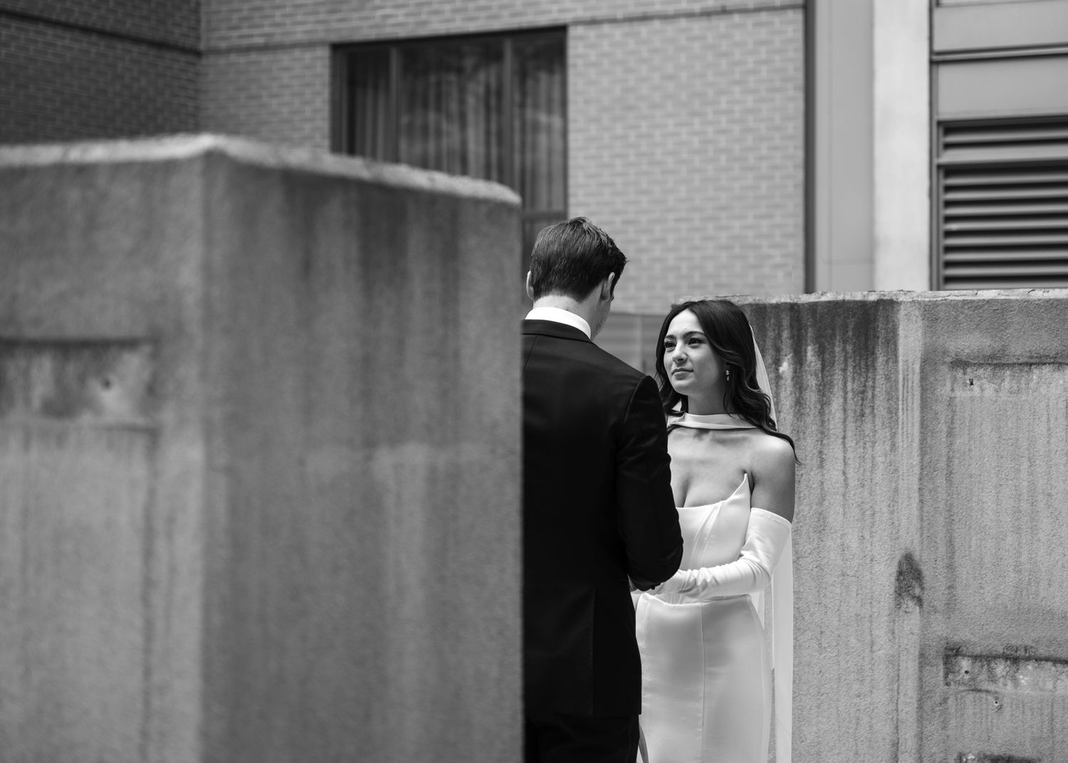A couple in wedding attire share an intimate moment against a brick wall backdrop in black and white.