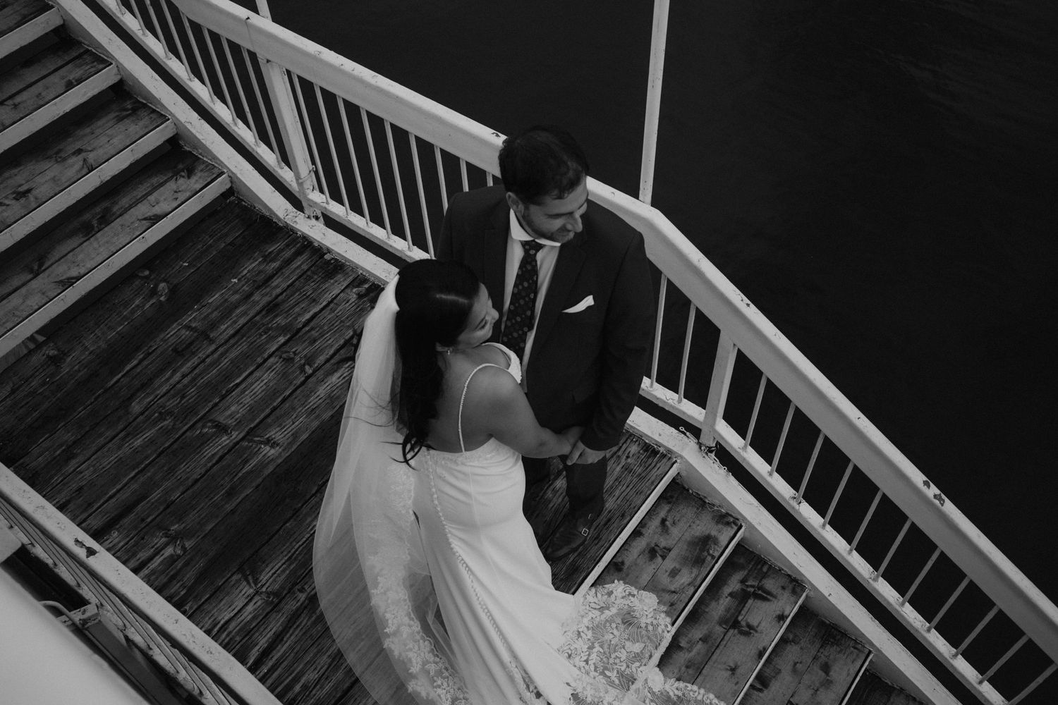 Black and white photograph of a couple in wedding attire on wooden deck stairs.