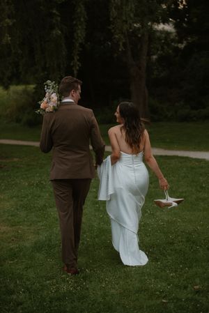 Newlyweds in brown suit and white dress walk hand in hand through a lush green garden.