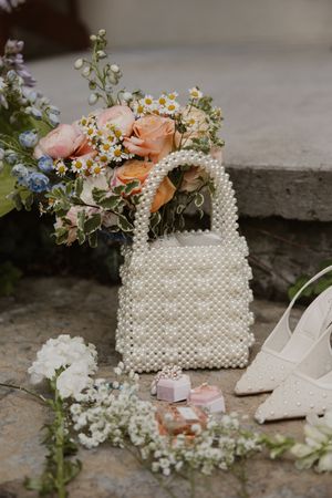 Delicate beaded handbag adorned with peach roses and baby's breath sits beside elegant white bridal shoes.