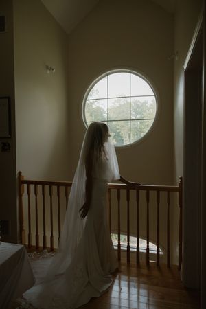 Ethereal silhouette stands near circular window in moody hallway with wooden banister.
