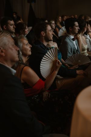 Theater audience members hold decorative fans while watching a performance in dim lighting.