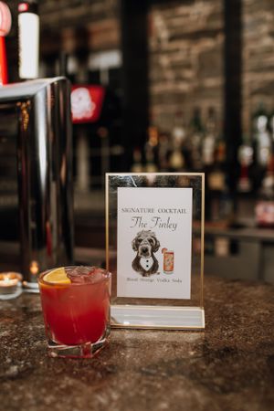 A red cocktail sits next to a signature cocktail menu card titled 'The Finley' on a bar counter.