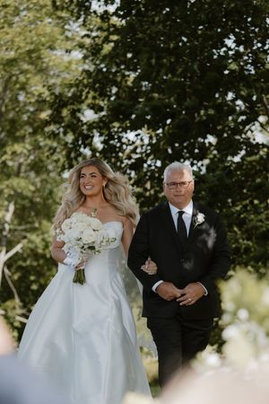 A bride in a flowing white gown walks arm-in-arm with her escort down an outdoor path surrounded by trees.