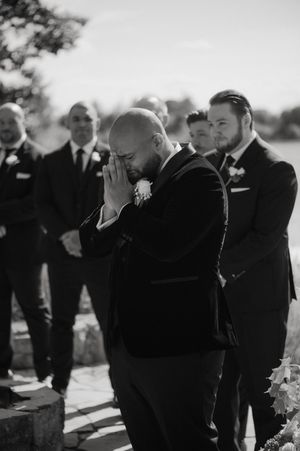 Black and white photo of an emotional moment at an outdoor wedding ceremony as a groom wipes tears while groomsmen look on.