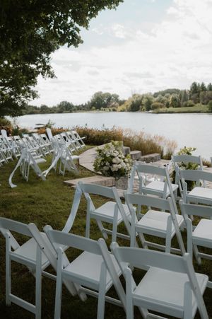 White folding chairs set up for an outdoor lakeside wedding ceremony with floral arrangements and water views.