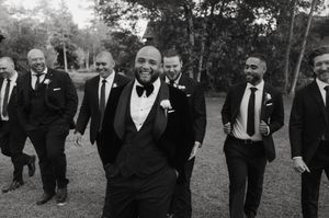 Groomsmen in black suits walk through a garden setting in a candid wedding photo.