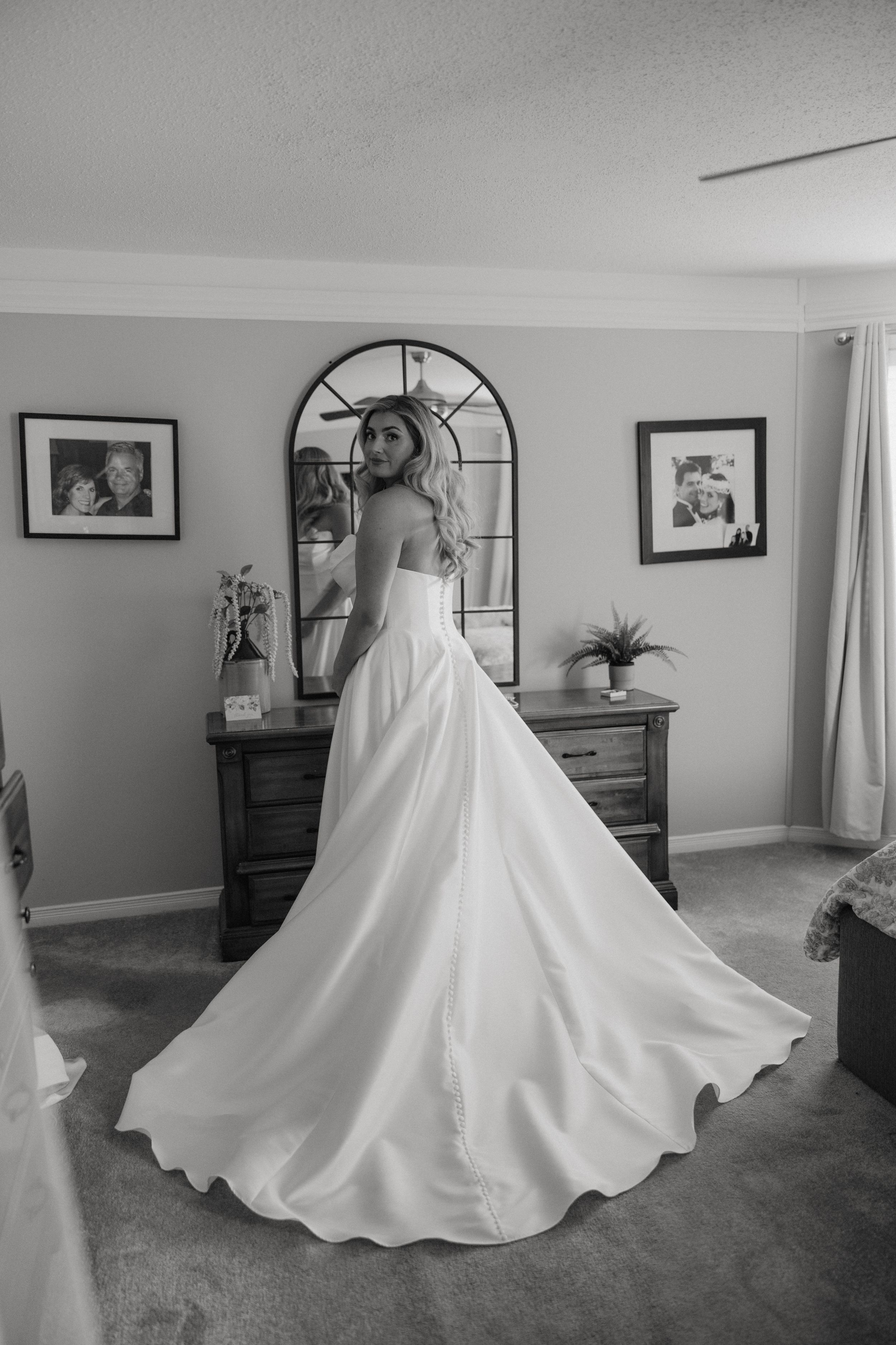 A stunning white wedding gown with a dramatic full skirt photographed in front of an arched mirror in a bedroom.