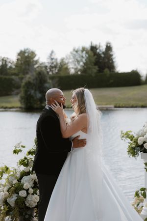 Romantic wedding couple shares intimate moments by a serene lake with white floral arrangements.