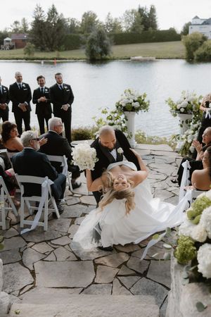 Wedding party and guests seated on stone patio overlooking lake during outdoor ceremony.