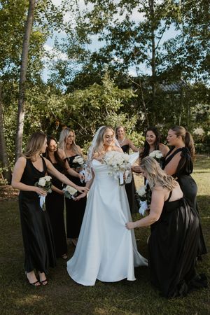 Wedding party in black dresses helps adjust the bride's white gown during an outdoor celebration.