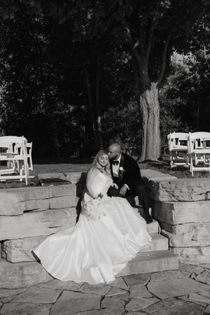 A black and white wedding photo of a couple sitting together on stone steps surrounded by trees.