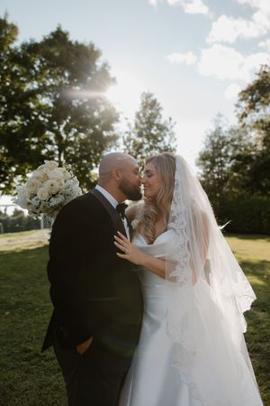 A romantic outdoor wedding moment captured in sunlight with a flowing white veil and bouquet of white flowers.