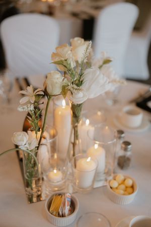 Elegant wedding reception table decor with white flowers in vases and glowing candles on white tablecloths.