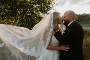 A romantic wedding couple embraces in a golden field during sunset as a veil flows dramatically in the breeze.