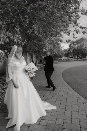 A bride in a flowing white wedding dress walks along a brick pathway under tree canopies in a garden setting.