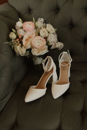 Elegant white bridal heels and soft pink rose bouquet arranged on dark tufted leather couch.