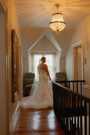 A bride in a flowing white wedding dress stands in an elegant hallway with dark wood railings and crystal chandeliers.