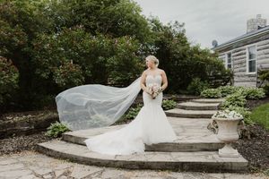 A flowing white wedding dress with a long veil catches the breeze on stone garden steps surrounded by greenery.