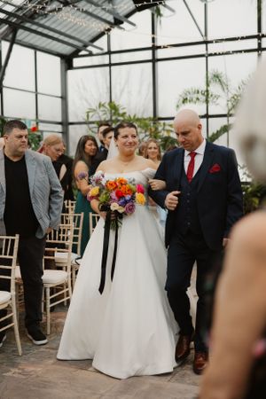 A wedding processional takes place inside a beautiful greenhouse filled with natural light and tropical greenery.