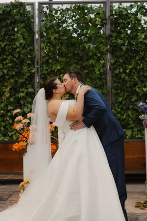 Romantic moment as newlyweds share a kiss in front of lush greenery and orange flowers in a greenhouse setting.