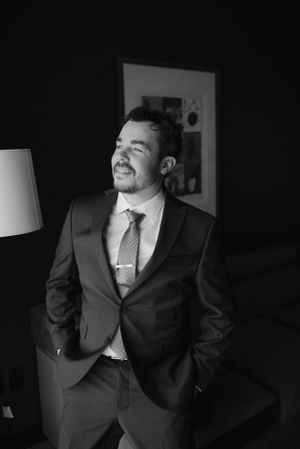 A black and white photo of someone in a formal suit smiling while standing in a dimly lit room.