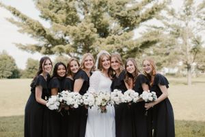 Bridal party in black and white dresses pose together holding white bouquets outdoors.