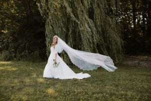 A bride in a flowing white wedding dress poses with her veil dramatically flowing in front of a weeping willow tree.