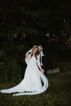 Elegant couple in wedding attire pose against greenery background.