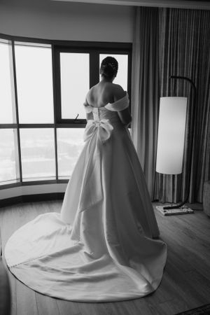 A bride in an elegant white ball gown with a dramatic bow detail stands by a window in a black and white photograph.