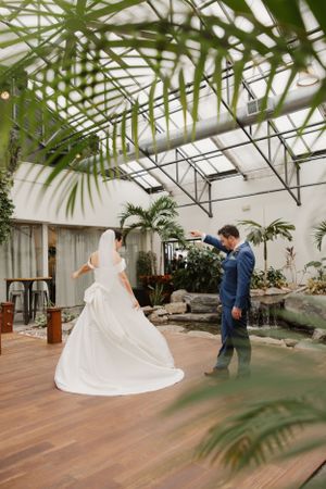 A first look wedding moment captured in a greenhouse venue with tropical plants and wooden flooring.